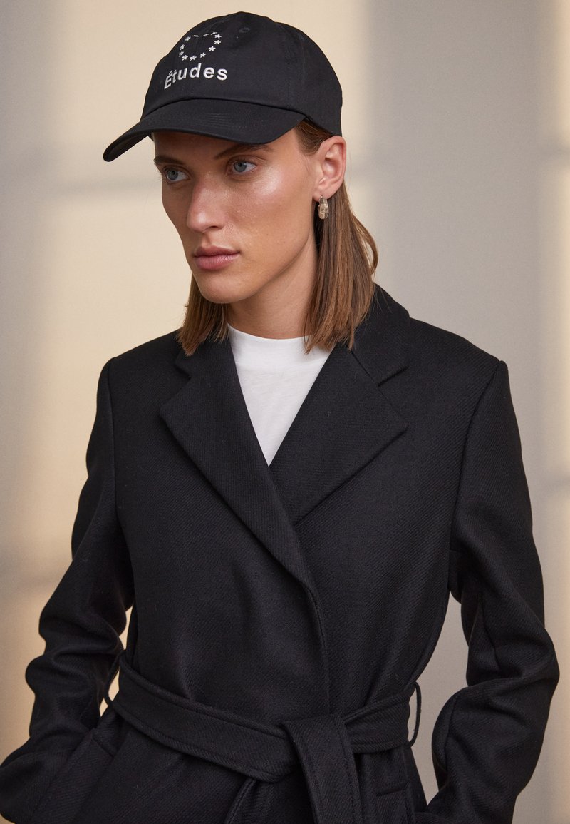 Black cap with "Études" logo, worn with a black coat featuring a double-breasted design and belted waist, layered over a white shirt.
