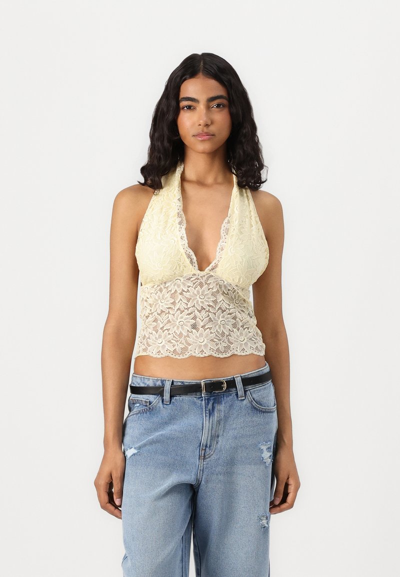 Čipkovaný crop top v svetložltej farbe s kvetinovým vzorom, hlbokým V-výstrihom a zakončením s volánikmi, skombinovaný s poškodenými modrými džinami a čiernym opaskom.