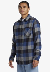 Man in een blauw, zwart en grijs geblokt overhemd met borstzak, lichte spijkerbroek, staande tegen een witte achtergrond.