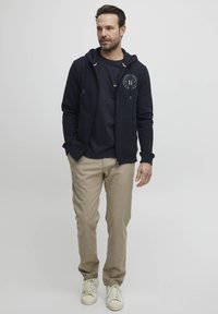 Haut à fermeture éclair marine avec logo, accompagné d'un t-shirt sombre et d'un pantalon beige clair. Les chaussures sont des baskets blanches. Tenue simple et décontractée.