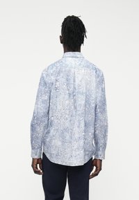 Šviesiai mėlyna ilgomis rankovėmis marškinėliai su gėlių raštu, sagų apykakle ir tekstūruota medžiaga. Nugara matomas sagstomi rankogaliai ir siūlės.