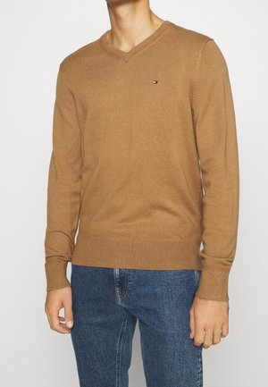 Mann trägt einen braunen V-Ausschnitt-Pullover mit kleinem Logo auf der Brust und blaue Jeans vor einfachem Hintergrund.