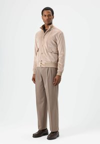 Beige suède bomberjack met drukknopen en twee frontzakken, gecombineerd met lichtbruine broek en donkere schoenen, staand op een effen achtergrond.