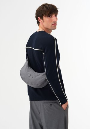 Grau, weicher Stoffbeutel, über den Rücken getragen, mit zwei runden metallen Akzenten; das Modell trägt einen marineblauen Pullover mit weißen Paspeln. Graue Hose.