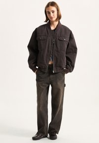 Dunkle, übergroße Bomberjacke mit zwei Vordertaschen, gerippten Bündchen und einem Reißverschluss. Kombiniert mit locker sitzenden dunklen Jeans und schwarzen Schuhen.