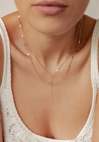 Collares en capas de oro rosa con cadenas finas, piezas de acento geométricas y un diseño de caída en forma de Y, reposando sobre una blusa blanca texturizada.