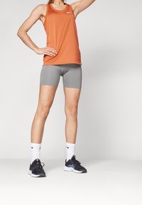 Persoon staat met één hand op de heup, gekleed in een oranje mouwloos topje, grijze fietsbroekjes, witte sokken en zwarte sportschoenen.
