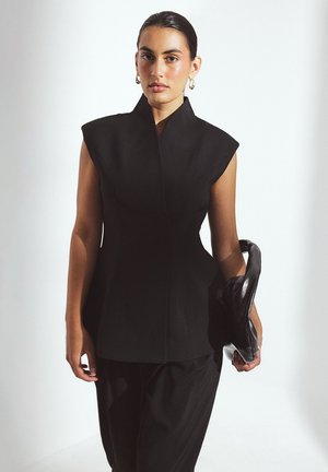 Top estructurado negro con cuello alto y mangas tipo capa, combinado con pantalones negros sueltos; tejido suave y diseño minimalista.