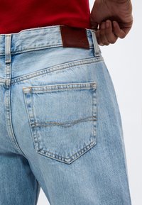 Φωτεινά γαλάζια τζιν με πίσω τσέπη και δερμάτινη ετικέτα επωνυμίας, φορεμένα από άτομο με κόκκινο τοπ που ρυθμίζει τη μέση του παντελονιού.