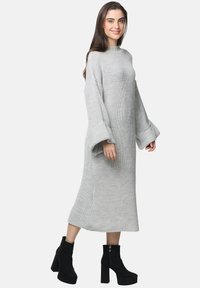 Hellgraues Strickkleid mit lockerer Passform, langen Ärmeln und ausgestellt geschnittenen Manschetten, kombiniert mit schwarzen Wildleder-Ankle-Boots. Glatte Textur, knöchellang.