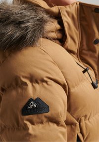 Veste matelassée beige avec une bordure de capuche en fausse fourrure, une texture matelassée et un patch logo noir avec un motif de montagne sur la manche.