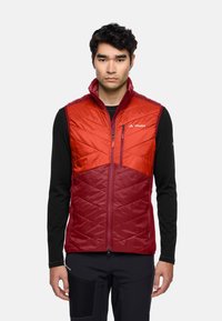 Gilet isolato rosso e marrone bordeaux con un design trapuntato, cerniera frontale e colletto rialzato. Indossato sopra una maglietta nera a maniche lunghe.