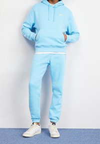 Person som bär en matchande ljusblå Nike-huvtröja och sweatpants med vita sneakers, stående mot en vit vägg och en blå matta.