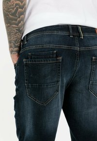 Mørkeblå denimjeans med to baglommer med skrå låg, synlige syninger og en tætsiddende pasform.