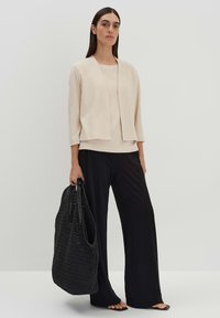 Beige strukturierte Strickjacke mit offenem Front, getragen über einem passenden Oberteil, kombiniert mit schwarzen weit geschnittenen Hosen und einer großen schwarzen geflochtenen Tragetasche.