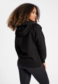 Felpa nera con cappuccio, dal fit rilassato e polsini a costine. Realizzata in tessuto morbido e presenta un'etichetta con logo vicino all'orlo, abbinata a leggings neri.