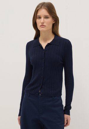 Femme portant un cardigan côtelé bleu marine à boutons avec manches longues et un pantalon bleu marine assorti, debout devant un fond clair uni.