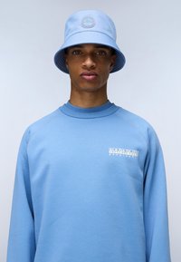 Felpa azzurra con collo alto e maniche raglan, caratterizzata da un logo bianco del marchio sul petto, abbinata a un cappello a secchiello blu coordinato.
