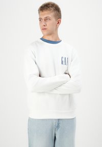 Sudadera blanca con cuello azul, que presenta un logo azul de "GAP" en el frente. Tejido suave con mangas largas y un corte relajado. Jeans de mezclilla de color azul claro.