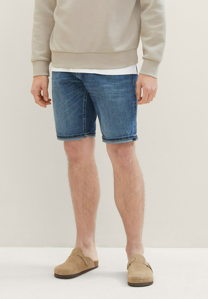 Jeansshorts in einem mittleren Blauton mit lockerer Passform, umgeschlagenem Saum und leichter Ausbleichung. Kombiniert mit einem hellbeigen Sweatshirt und Slippern.