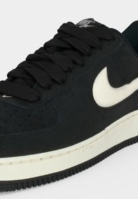 Μαύρο αθλητικό παπούτσι Nike με λευκό λογότυπο Swoosh, μαύρα κορδόνια και λευκή σόλα με μαύρο κάτω μέρος, εμφανίζεται από μπροστινή πλευρική γωνία.