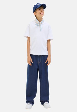 Child smiles while wearing a white t-shirt, blue scarf, navy cap, and loose blue jeans, standing against a plain background.