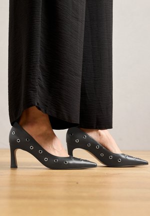 Scarpe col tacco alto in pelle nera con design a punta, caratterizzate da dettagli in metallo lungo i lati e un sottile tacco a stiletto.