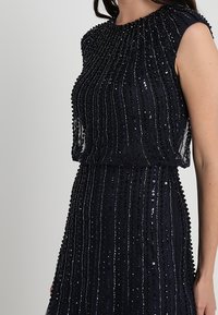 Vestido sin mangas azul marino con una superposición texturizada, adornado con rayas verticales de lentejuelas y cuentas. Cuello redondo, cintura ceñida.