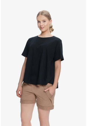 Kobieta ubrana w luźną czarną koszulkę z krótkim rękawem i beżowe szorty z mankietami, stojąca z jedną ręką w kieszeni na jednolitym białym tle.