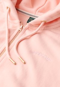 Sudadera con cremallera de color rosa claro, hecha de un tejido suave. Presenta herrajes en tono dorado, un logotipo bordado "Superdry" y cordones ajustables.