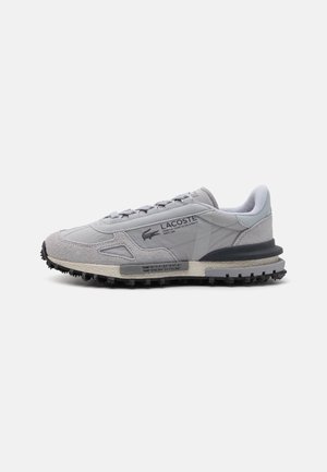 Zapatilla deportiva gris con parte superior de ante y textil, que presenta una suela de goma texturizada, cierre de cordones y el logo de Lacoste en el lateral.