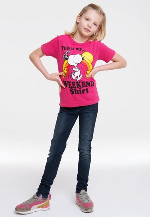 Rosa kurzärmeliges T-Shirt mit Cartoon-Charakteren und Text, kombiniert mit dunkelblauen Jeans und mehrfarbigen Turnschuhen.