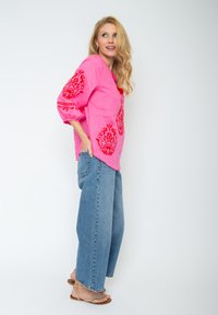 Rosa Leinenbluse mit roter Blumenstickerei, Drei-Viertel-Ärmel und lockerer Passform, kombiniert mit hellblauen weit geschnittenen Jeans.