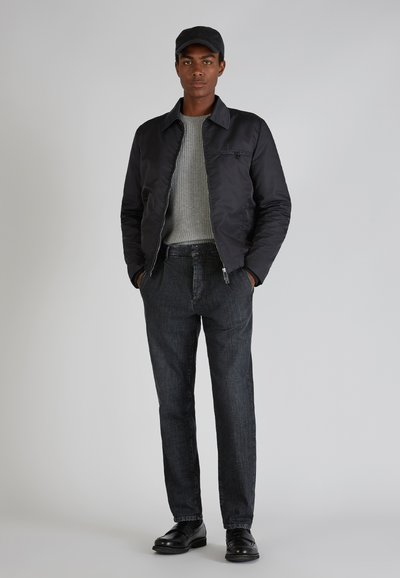 Giacca bomber nera, maglione grigio a maglia, pantaloni in denim scuri, scarpe nere e cappellino nero. La giacca ha una chiusura con zip e tasche laterali.