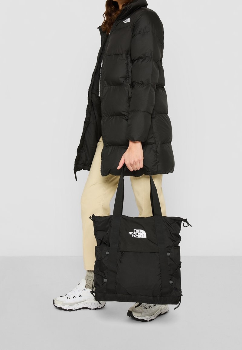 Bolso tote negro con doble asa, hecho de tela texturizada, cuenta con un bolsillo frontal y acentos de cordón ajustable. Logotipo visible.