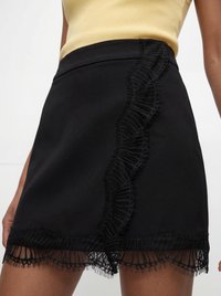 Gros plan d'une mini-jupe noire avec un bord froncé vertical et un ourlet en dentelle portée avec un top jaune sur fond blanc.