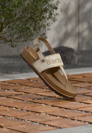 Șlapi de culoare bronz cu o bretea lată din material textil cu logo-ul „Barb”, talpă texturată și o cataramă ajustabilă la gleznă, așezați pe o suprafață din lemn.