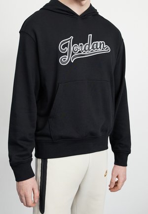 Person trägt einen schwarzen Kapuzenpullover mit dem weißen Schriftzug "Jordan" und hellfarbige Hosen mit einem kleinen Nike-Logo am Oberschenkel.