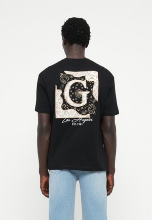 Camiseta negra con un gran gráfico que presenta una letra 'G' estilizada en color crema y dorado, con intrincados patrones de paisley y florales, y "Los Ángeles EST. 1981."
