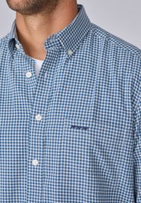 Chemise à carreaux bleue et blanche avec col, dotée d'une poche avant et d'une broderie subtile. Fabriquée en tissu léger.