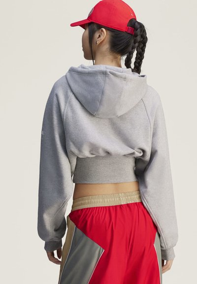 Mujer con el cabello trenzado lleva una sudadera corta gris con capucha, pantalones rojos con bloques de color y una gorra de béisbol roja, de espaldas a la cámara.