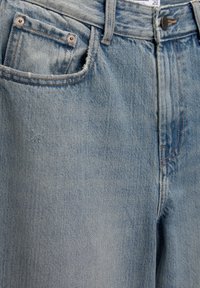 Jean en denim bleu clair avec poches avant, coutures visibles, rivets métalliques et fermeture par bouton à la taille.