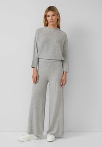 Femme se tenant contre un mur gris, portant un haut en tricot gris clair assorti et un pantalon large, avec des baskets blanches.