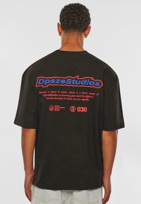 Schwarzes Baumwoll-T-Shirt mit kräftigen blauen und roten Grafiken auf der Rückseite. Der Text enthält "DpszeStudios" und ein motivierendes Zitat, ergänzt durch Logos.