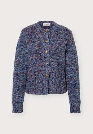Femme debout portant un cardigan en tricot bleu multicolore avec des boutons, un pantalon large noir et des chaussures noires sur un fond gris clair.