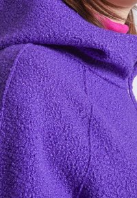 Sweat à capuche en polaire violet avec une surface texturée, doté d'une capuche et de détails de coutures visibles, porté par-dessus un vêtement rose.
