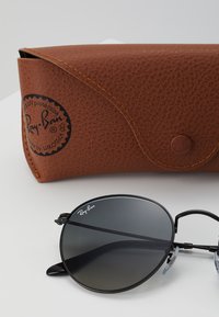 Ochelari de soare Ray-Ban cu lentile circulare negre și ramă subțire din metal negru. Însoțiți de un toc din piele texturată maro, cu logo.