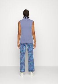 Ärmlös, ljuslila topp med hög krage, tillsammans med vida blå byxor med en färgglad ögonmönstrad design. Vita skor.