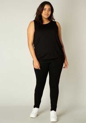 Vrouw staand, gekleed in een zwart mouwloos topje, zwarte legging en witte sneakers, tegen een effen lichte achtergrond.