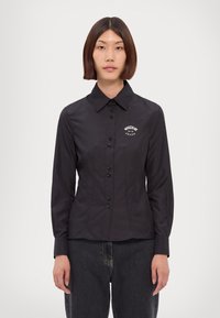 Donna con capelli castani lisci indossa una camicia nera attillata con il logo Moschino Jeans sul petto e jeans scuri, in piedi contro uno sfondo bianco.